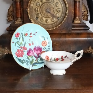 May include: A light blue saucer featuring a colorful floral pattern with pink, red, and orange flowers, paired with a white teacup that has a matching floral design and a gold-trimmed rim, both resting in front of an antique wooden clock.