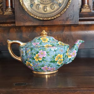 Könnte beinhalten: Eine Porzellan-Teekanne mit hellblauem Hintergrund und einem dichten Muster aus gelben und rosa Blumen sowie grünen Blättern. Die Teekanne hat einen passenden Deckel mit goldenem Knauf, einen goldverzierten Henkel, Ausguss und Boden.