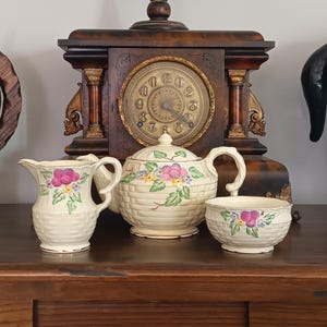 Puede incluir: Un juego de té de cerámica color crema que consta de una tetera, una lechera y un azucarero. Cada pieza presenta una textura de tejido de cesta moldeada y está decorada con racimos de flores moradas, hojas verdes y pequeños detalles azules.