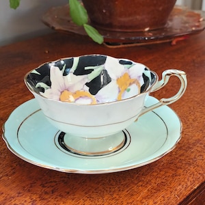 Puede incluir: Un juego de taza y platillo vintage con un diseño floral. La taza tiene un patrón floral blanco y negro con detalles dorados. El platillo es de color azul claro con un borde dorado.