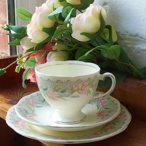 May include: A vintage cream-colored teacup, saucer, and plate set with a floral design. The set features pink flowers and green leaves, with gold trim. A bouquet of pink and white roses sits behind the teacup on a wooden surface.