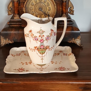 Puede incluir: Un juego de jarra y bandeja vintage de color crema. La jarra presenta un diseño floral con detalles en rosa, naranja y verde. La bandeja tiene un borde festoneado y un patrón floral similar. Un reloj decorativo está en el fondo.