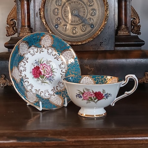 Puede incluir: Un juego de taza y platillo de té vintage. La taza y el platillo blancos presentan un diseño floral con flores rosas y rojas, detalles azules y ribetes dorados. El platillo tiene un borde turquesa con filigrana dorada.