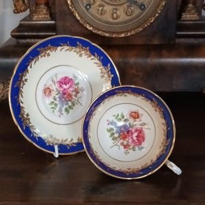 Puede incluir: Juego de taza y platillo de té vintage. La taza y el platillo son de color crema con un borde azul y ribetes dorados. Un diseño de ramo floral está en el centro. Se muestra sobre una superficie de madera oscura.