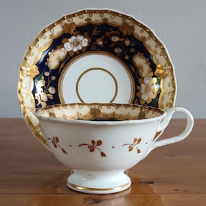 May include: A white teacup and saucer set with gold and floral accents. The saucer has a navy blue center with gold trim and floral designs. The teacup features gold floral sprigs and a gold rim. The set is on a wooden surface.