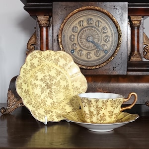 May include: A vintage yellow teacup, saucer, and plate set with a gold floral design. The teacup has a curved handle and sits on a matching saucer. The plate is scalloped. The set is displayed in front of a dark wooden clock.