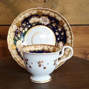 Puede incluir: Juego de taza y platillo de té blanco con detalles florales dorados. El platillo tiene un borde azul marino con detalles dorados. La taza de té presenta un borde dorado y una pequeña base con pie. El juego se exhibe sobre una superficie de madera oscura.