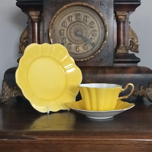 May include: A yellow teacup and saucer set with a matching plate. The teacup and plate have a scalloped edge design. The teacup has a gold handle and trim. The set is on a wooden surface, with a vintage clock in the background.