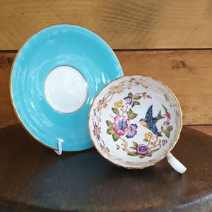 May include: A vintage teacup and saucer set. The saucer is a bright turquoise with a gold rim. The teacup is white with a floral and bird design, also with a gold rim. The set is displayed on a dark brown surface.