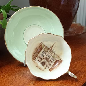 Puede incluir: Un juego de taza y platillo de té vintage. El platillo es de color verde claro con un borde dorado, y la taza presenta una ilustración arquitectónica detallada con el texto "Greifswald".