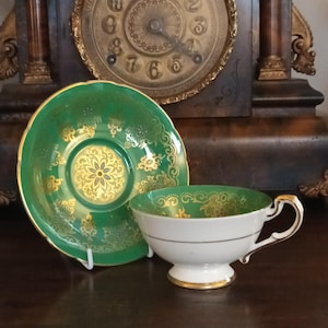 Puede incluir: Un juego de taza y platillo de té vintage. La taza es blanca con ribetes dorados y un asa dorada. El platillo y el interior de la taza son verdes con diseños florales dorados. El juego se exhibe frente a un reloj de madera oscura.
