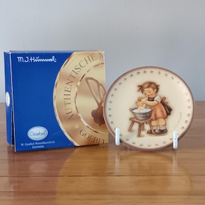 May include: A decorative plate depicting a girl washing a teddy bear, with a star-patterned border. The plate is displayed on a white stand beside a blue box with "m.j. hummel" and "Goebel" printed on it.
