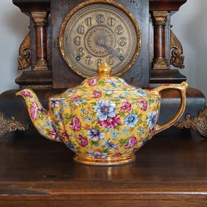 Könnte beinhalten: Eine gelbe Teekanne mit Blumenmuster und goldenem Griff, Ausguss und Boden. Die Teekanne hat einen Deckel mit goldener Spitze und ist mit rosa, blauen und lila Blumen verziert. Die Teekanne steht auf einer Holzoberfläche vor einer Vintage-Uhr.