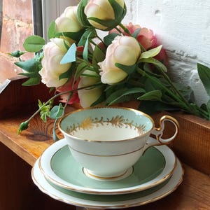 Peut inclure: Service à thé vintage comprenant une tasse, une soucoupe et une assiette. La tasse a un bord vert menthe avec des détails floraux dorés et une anse dorée. La soucoupe et l'assiette sont vert menthe avec un bord blanc. Un bouquet de roses est derrière.