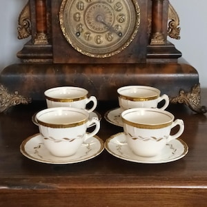 May include: Four white teacups and saucers with gold trim are displayed on a wooden surface. The teacups have a simple design, and the saucers have a matching gold pattern. A vintage clock with a gold-coloured face is in the background.