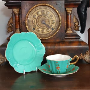 May include: A teal ceramic teacup and saucer set featuring a scalloped edge with gold trim and orange floral accents, accompanied by a matching teal scalloped saucer standing upright, all displayed in front of a dark wood mantle clock.