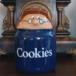 Pode incluir: Um pote de biscoitos de cerâmica azul com uma tampa em forma de rosto de desenho animado. A tampa apresenta um personagem com cabelo castanho e óculos. O pote é cilíndrico com a palavra "Cookies" em letras brancas.