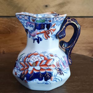 Può includere: Piccolo brocca in ceramica bianca con manico blu e design floreale. La brocca presenta un motivo dettagliato di fiori arancioni e blu con accenti dorati. Il bordo ha un bordo decorativo blu e rosa.