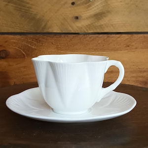 Può includere: Un set di tazza e piattino da tè bianchi. La tazza ha un manico curvo e un design scanalato. Il piattino è rotondo con un motivo scanalato abbinato. Il set è in porcellana e si trova su una superficie di legno scuro.