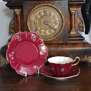 May include: A deep red ceramic tea cup and saucer set with a scalloped edge, gold trim, and small blue floral accents. A matching red plate with similar floral patterns stands upright next to the cup, all positioned in front of a wooden clock.