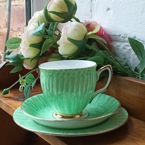 Pode incluir: Conjunto de chá verde vintage com chávena, pires e prato lateral com acabamentos dourados. A chávena e o pires apresentam um design texturizado. Flores artificiais no fundo.