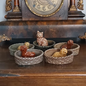 Może przedstawiać: Kolekcja ceramicznych figurek kotów i psów w koszach w stylu plecionym. Figurki są w różnych kolorach, w tym brązowym, beżowym i szarym. Kosze są w odcieniach brązu i szarości. Z tyłu znajduje się zabytkowy zegar.