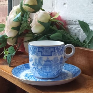 May include: A blue and white teacup and saucer set with a gold rim. The cup and saucer feature a detailed landscape design. The set is placed on a wooden surface, with a bouquet of flowers in the background.
