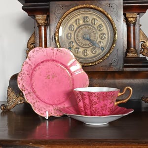 May include: A pink teacup and saucer set with gold detailing and a matching plate. The teacup and plate feature a floral design. The set is placed in front of a vintage clock.