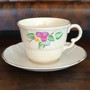 Puede incluir: Un juego de taza de té y platillo de porcelana color crema con un patrón texturizado de tejido de cesta y un motivo floral pintado a mano en tonos morado, verde, amarillo y azul, acabado con un delicado borde dorado.