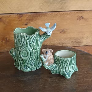 Può includere: Due fioriere in ceramica a forma di ceppo d'albero. Una è più alta con un uccello appollaiato sul lato. L'altra ha un koala seduto sul lato. Entrambe sono verdi con una trama di venature del legno.