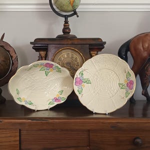 Puede incluir: Dos platos de cerámica de color crema con bordes festoneados y ribete dorado. Ambas piezas presentan un patrón texturizado de tejido de cesta en el centro y están decoradas con racimos pintados a mano de flores rosas, amarillas y azules.