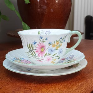 May include: A white teacup and saucer set with a floral pattern in shades of pink, blue, and yellow. The set has a green trim around the edges.