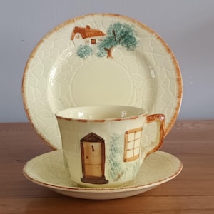 Può includere: Un servizio da tè vintage composto da una tazza, un piattino e un piatto. I pezzi in ceramica color crema presentano un design a casetta con una porta, una finestra e un tetto marroni. Il piatto e il piattino hanno un motivo strutturato e bordi marroni.