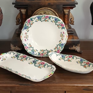 May include: A set of three cream-colored ceramic dishes featuring a scalloped edge and a decorative border of colorful flowers, green vines, and a lattice pattern. The collection includes one round bowl, one rectangular tray, and one oval dish.