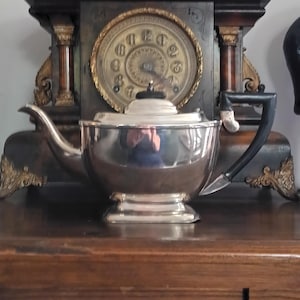 May include: A shiny silver teapot with a black handle and lid sits on a wooden surface. The teapot is in front of a vintage clock with a gold-colored face and Roman numerals. The clock is housed in a dark wooden frame.