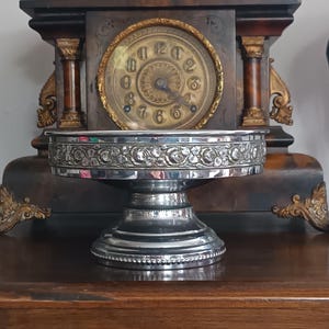 Puede incluir: Un cuenco de pedestal de metal plateado con un recipiente ancho y poco profundo que presenta una banda floral en relieve decorativa alrededor del borde exterior. El cuenco se apoya en una base circular resistente con un detalle de borde perlado.
