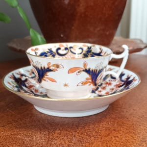 Puede incluir: Una taza de té y un platillo blancos con un patrón floral azul y naranja. La taza tiene un borde dorado y el platillo tiene una banda dorada alrededor del borde.