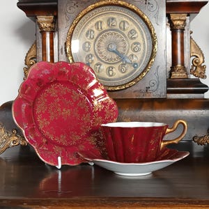 May include: A red teacup and saucer set with gold detailing and floral patterns. The teacup features a fluted design and a gold handle. A matching plate with a scalloped edge is also present. The set is displayed in front of an antique clock.