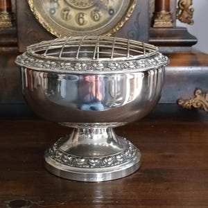 May include: A silver-colored, footed bowl with a grid insert, likely for floral arrangements. The bowl has a rounded body and a decorative, floral-patterned base. The background includes a vintage clock.