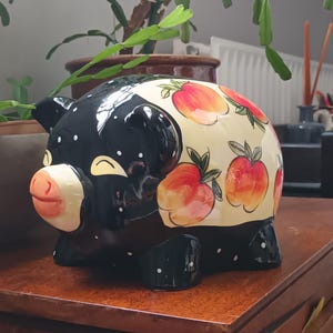 May include: A ceramic piggy bank with a black body and white polka dots. The top is cream-colored and decorated with watercolor-style peaches. The pig has a pink snout and ears. A decorative item for a home.