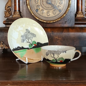 Könnte beinhalten: Ein Vintage-Tee-Set, bestehend aus einer Tasse und einer Untertasse. Die cremefarbene Tasse und Untertasse zeigen ein Landschaftsdesign mit einem Baum, grünen Büschen und einem Haus mit rotem Dach. Das Set wird auf einer dunklen Holzoberfläche mit einer Vintage-Uhr im Hintergrund präsentiert.
