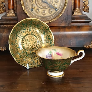 May include: A porcelain teacup and matching saucer featuring a deep green exterior with an intricate gold filigree pattern. The interior of the teacup displays a painted illustration of assorted fruits, including a pear, plum, and cherry.