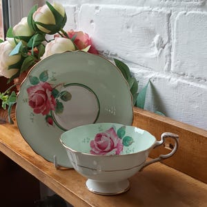 Puede incluir: Juego de taza y platillo de té vintage. La taza y el platillo son de color verde claro con un diseño de rosa rosa. La taza tiene una base blanca y ribete dorado. El platillo tiene un borde festoneado.