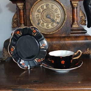 May include: A black porcelain tea cup and saucer set with a scalloped edge, featuring orange floral accents and gold trim. An additional matching black saucer with a scalloped edge and orange floral details stands upright next to the tea cup set.