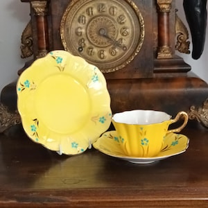 May include: A bright yellow tea cup and saucer set with a matching yellow side plate, all featuring a scalloped edge, gold trim, and small blue floral accents. The pieces have a ribbed texture and are displayed together on a flat surface.