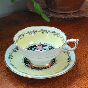 Puede incluir: Una taza de té y un platillo amarillos con un borde negro y dorado. La taza y el platillo tienen un diseño floral con flores rosas, blancas y verdes. La taza tiene una cinta negra con campanas doradas alrededor del borde.