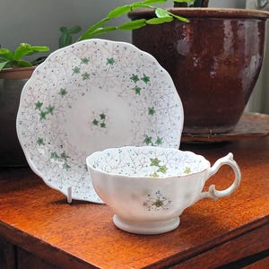Pode incluir: Um conjunto de chá branco com pires e xícara com um padrão de folhas verdes. A xícara tem uma alça curva e fica em um pé pequeno. O pires tem uma borda ondulada. O conjunto está em uma superfície de madeira.
