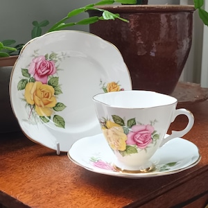 Pode incluir: Conjunto de chá vintage com chávena, pires e prato brancos. Decorado com rosas amarelas e rosas e folhas verdes. As bordas da chávena, pires e prato são douradas.