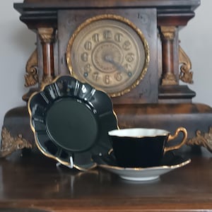 May include: A black teacup and saucer set with gold edging and a matching plate. The teacup has a curved handle and sits on a white-edged saucer. The plate has a scalloped edge. A vintage clock is in the background.
