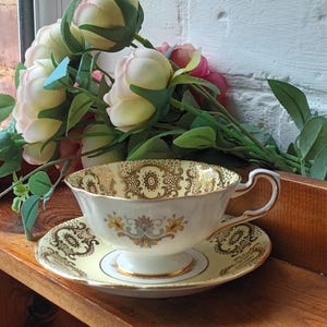 Puede incluir: Juego de taza y platillo de té vintage crema y amarillo con diseños florales y dorados. La taza se apoya sobre una base de pedestal. Flores artificiales en el fondo. El conjunto se exhibe en una estantería de madera.